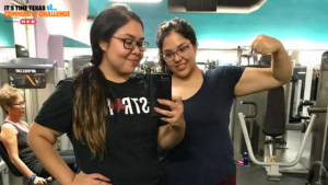 Two teenage girls are taking a photo in front of a mirror at the gym. One is showing off her arm muscles and the other has a hand on her hip.