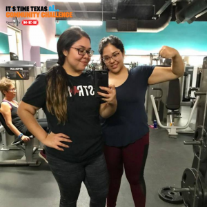 Two teenage girls in black workout clothes stand in front of the mirror at a gym taking a selfie. one is flexing the muscles in her arms and the other has a hand on her hips.