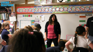 A female teacher in a pink shirt stands in front of the classroom at a whiteboard. She is smiling and facing her students who are all standing.