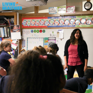 A teacher stands in front of her class. She is in front of a decorated whiteboard and is facing her students who are all standing and looking at her expectantly.