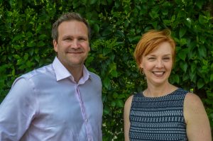 Amy McGeady, new CEO of It's Time Texas, wears a dark top and stands in front a wall of greenery. Baker Harrell, founder stands next to her. Both are smiling at the camera.