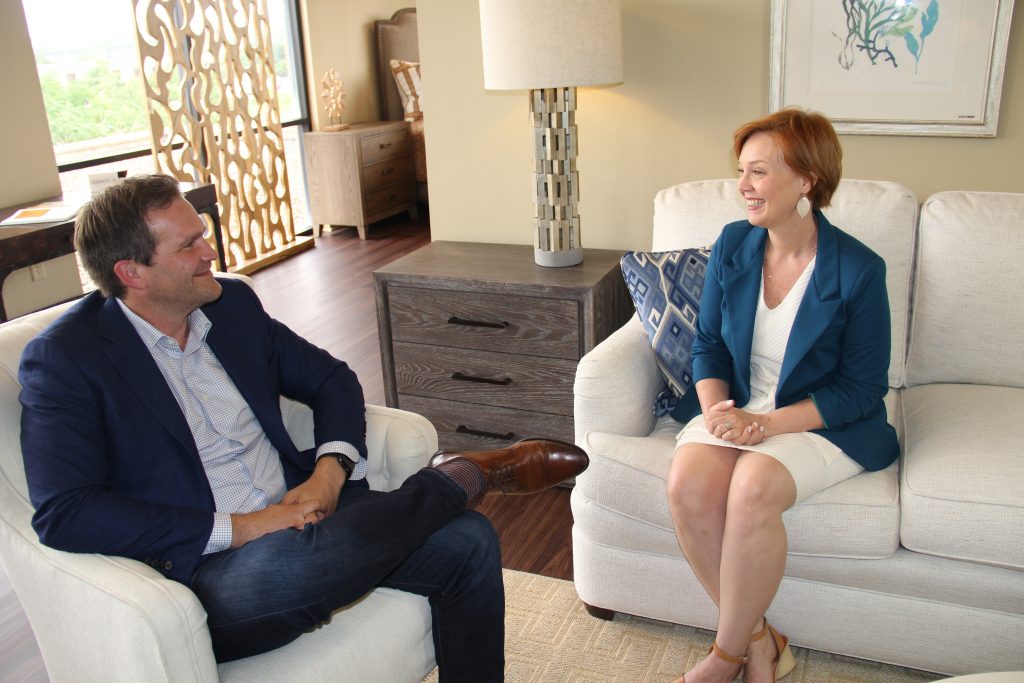 Amy McGeady, new CEO, and Baker Harrell, founder sit in a light and bright living room. McGeady sits on a couch, wearing a blue blazer and is smiling at Harrell. Harrell sits in a nearby armchair and is smiling at McGeady. 