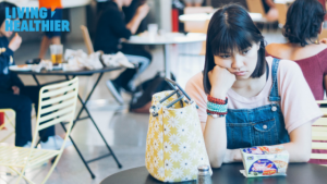 A teenage girl in overalls sits alone at a lunch table. Her head is in her hand and she is glumly looking at her school lunch.