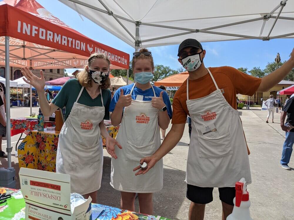 Three adults wearing Galveston's Own Farmers Market aprons at a local farmers market