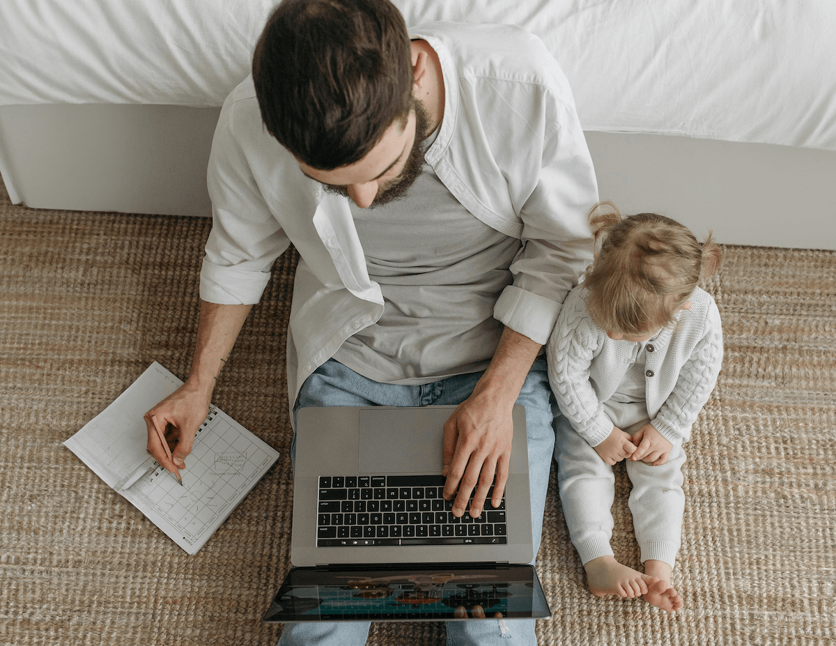 A father sitting next to his toddler-aged daughter while he works at home.